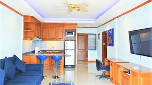 A modern kitchen with wooden cabinets, a microwave, and a stainless steel refrigerator. There is a blue sofa and a TV mounted on the wall.