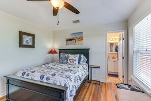 Un dormitorio con cama, ventilador de techo, mesita de noche, ventana con persianas, puerta que da a un baño y un cuadro en la pared.