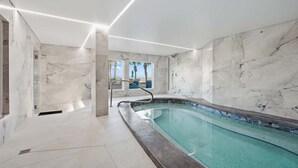 Une piscine intérieure moderne avec des murs et un plafond en marbre, une paroi en verre et une vue sur des palmiers et une plage.