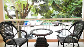 Un balcon avec des fauteuils en osier et une petite table ronde, donnant sur une rue bordée de voitures garées et d’arbres.