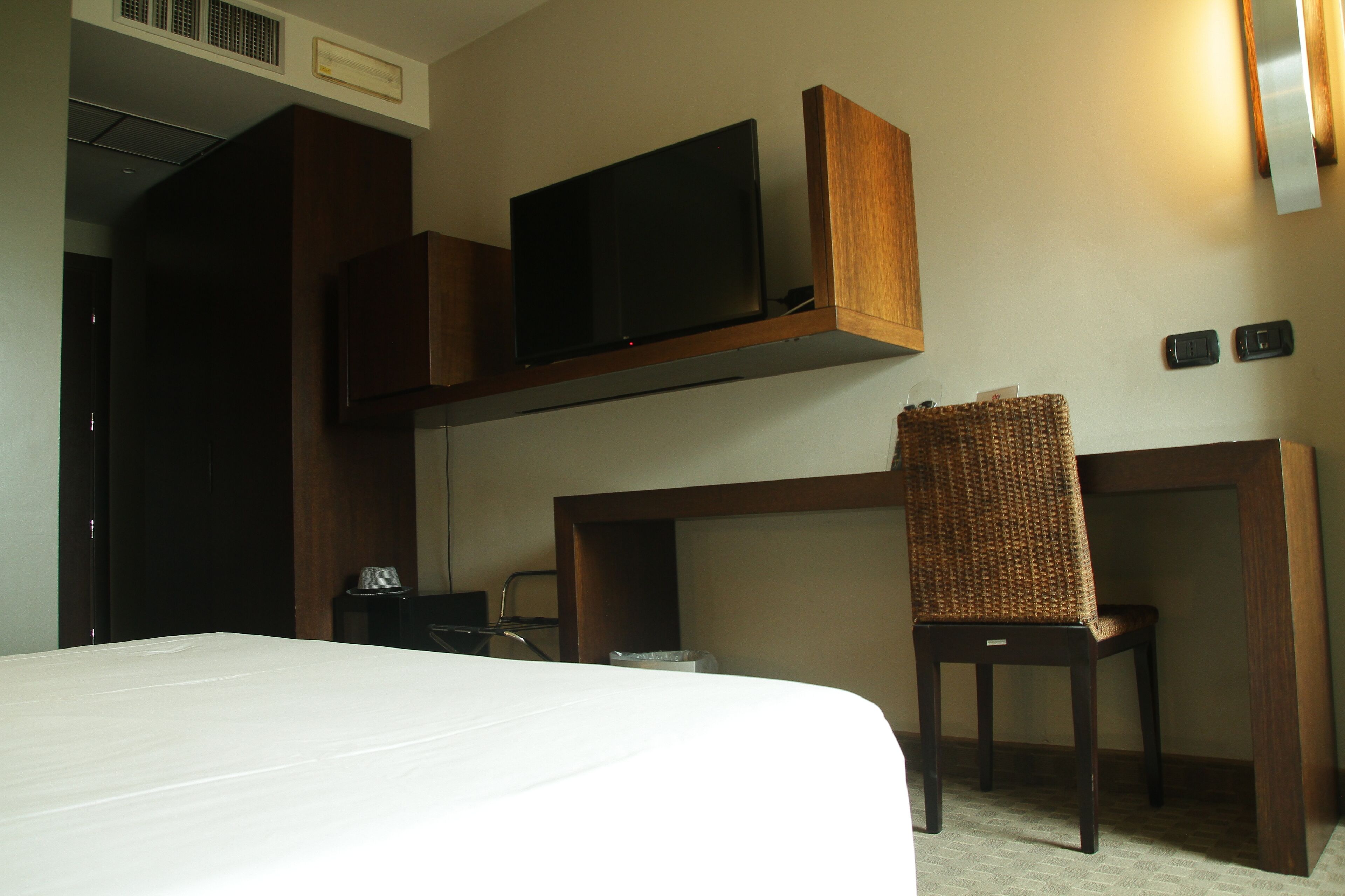 Habitación de hotel con cama, televisor en un estante de madera, escritorio con silla y una mesita de noche con lámpara.
