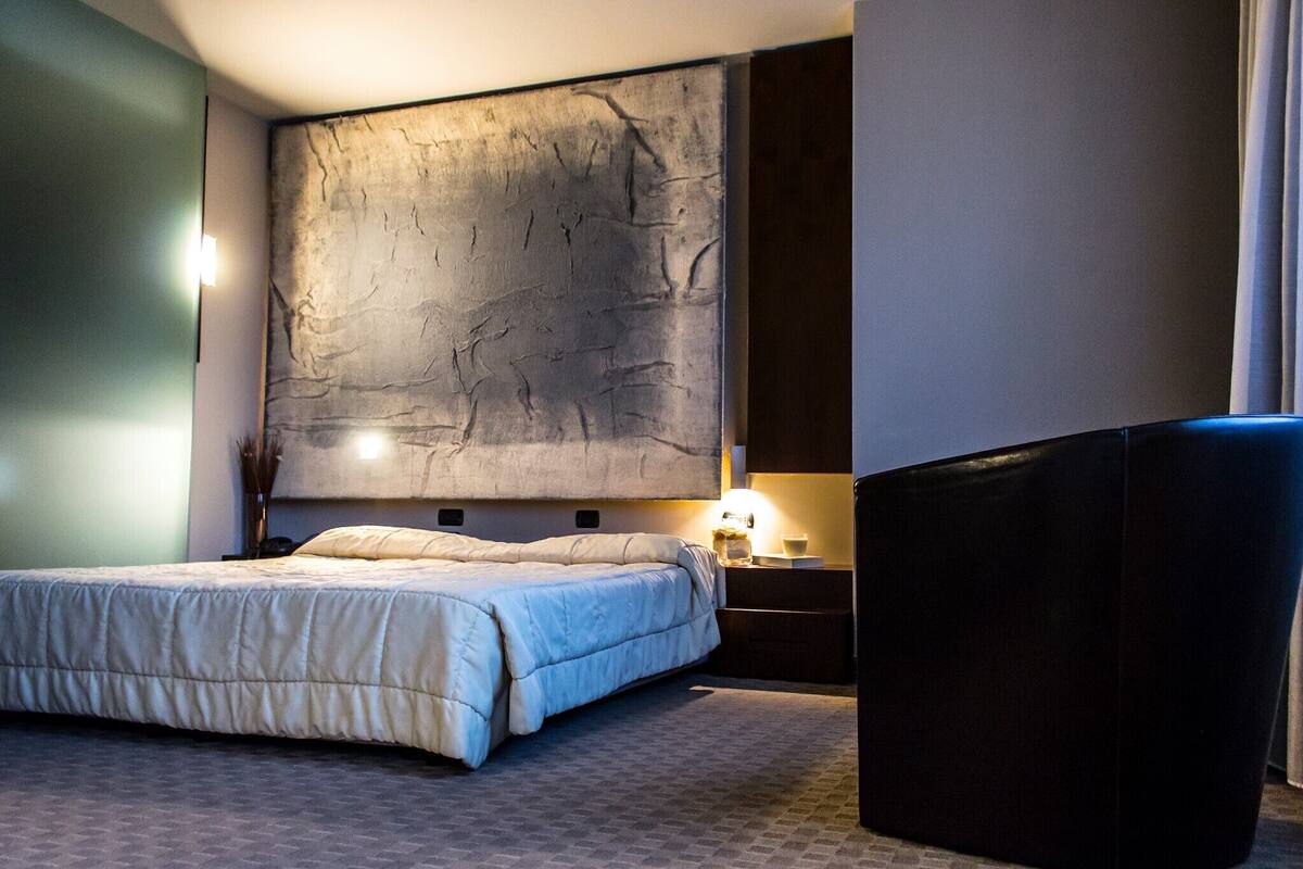 Habitació d'hotel amb un llit gran, una tauleta de nit amb una làmpada i una obra d'art moderna a la paret.