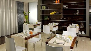 Un comedor moderno con sillas y mesas blancas, una estantería de madera oscura con objetos decorativos y un amplio ventanal con cortinas.