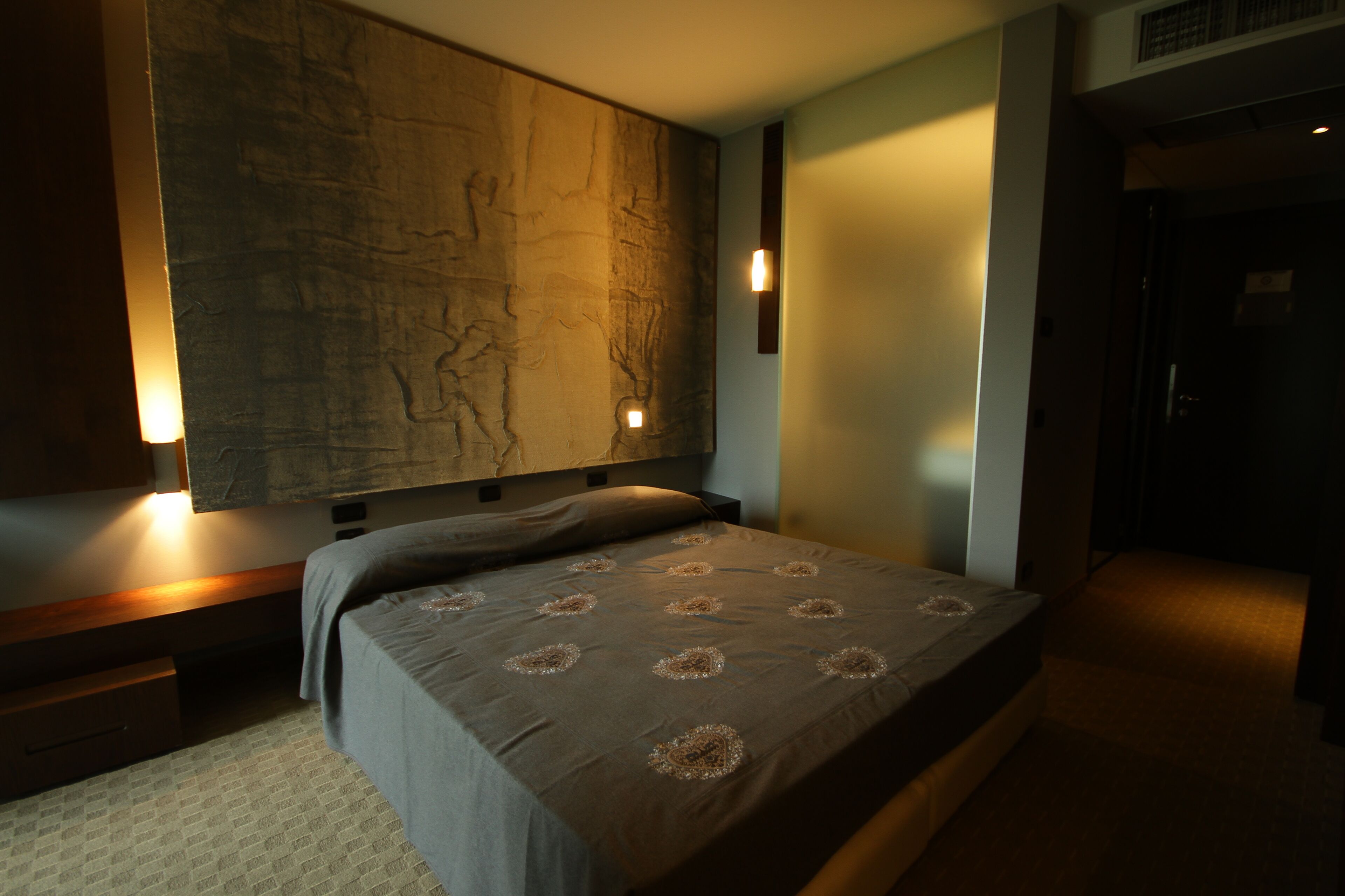 A bedroom with a large bed, a nightstand, a wall-mounted light, and a door.