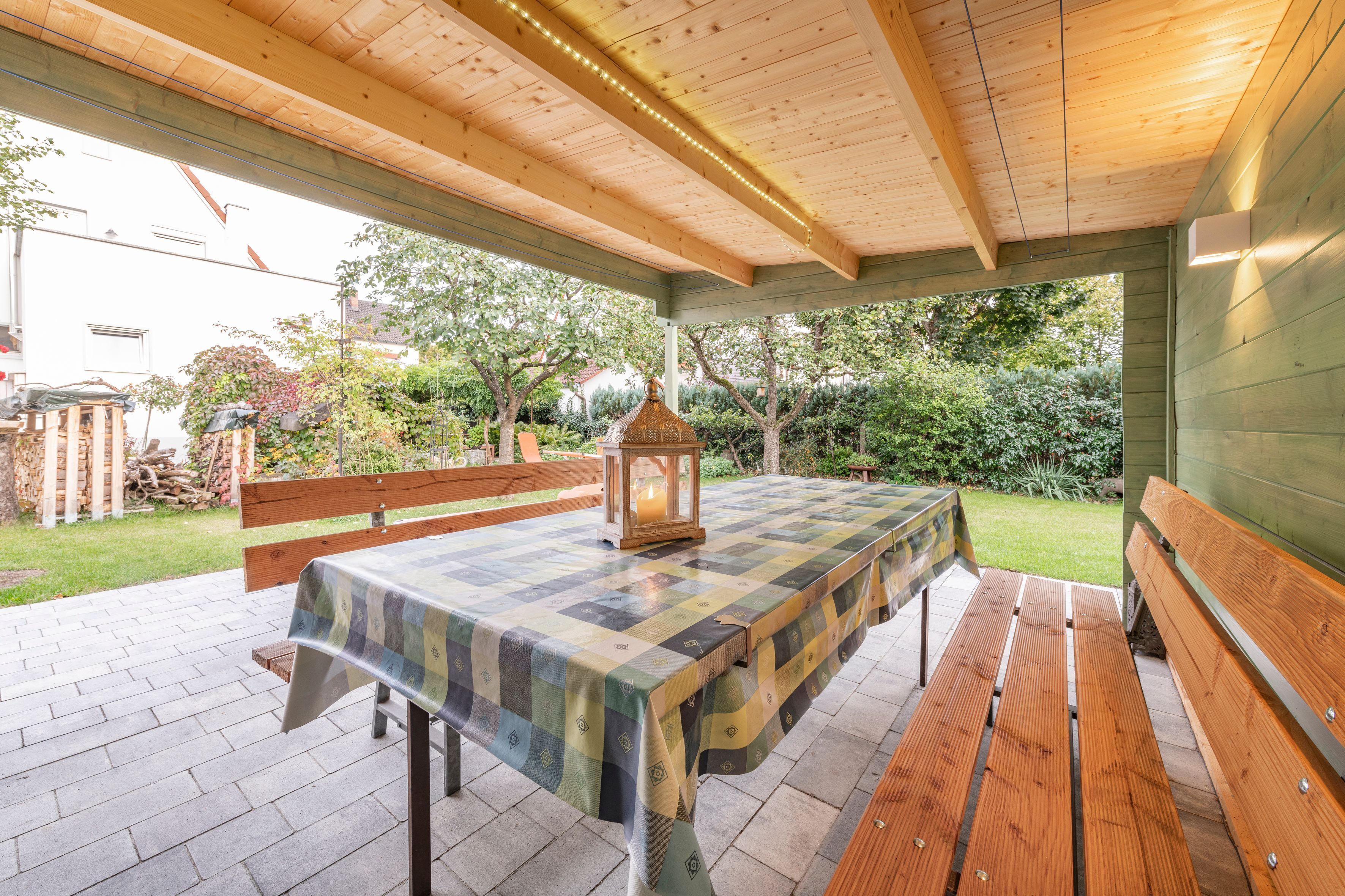 An outdoor patio with a wooden table and benches, surrounded by a garden.