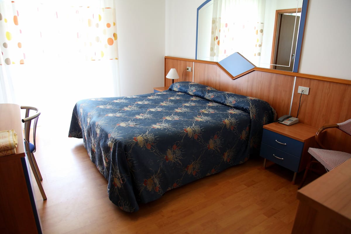 Un dormitorio con cama, mesita de noche, lámpara, silla y escritorio con teléfono.