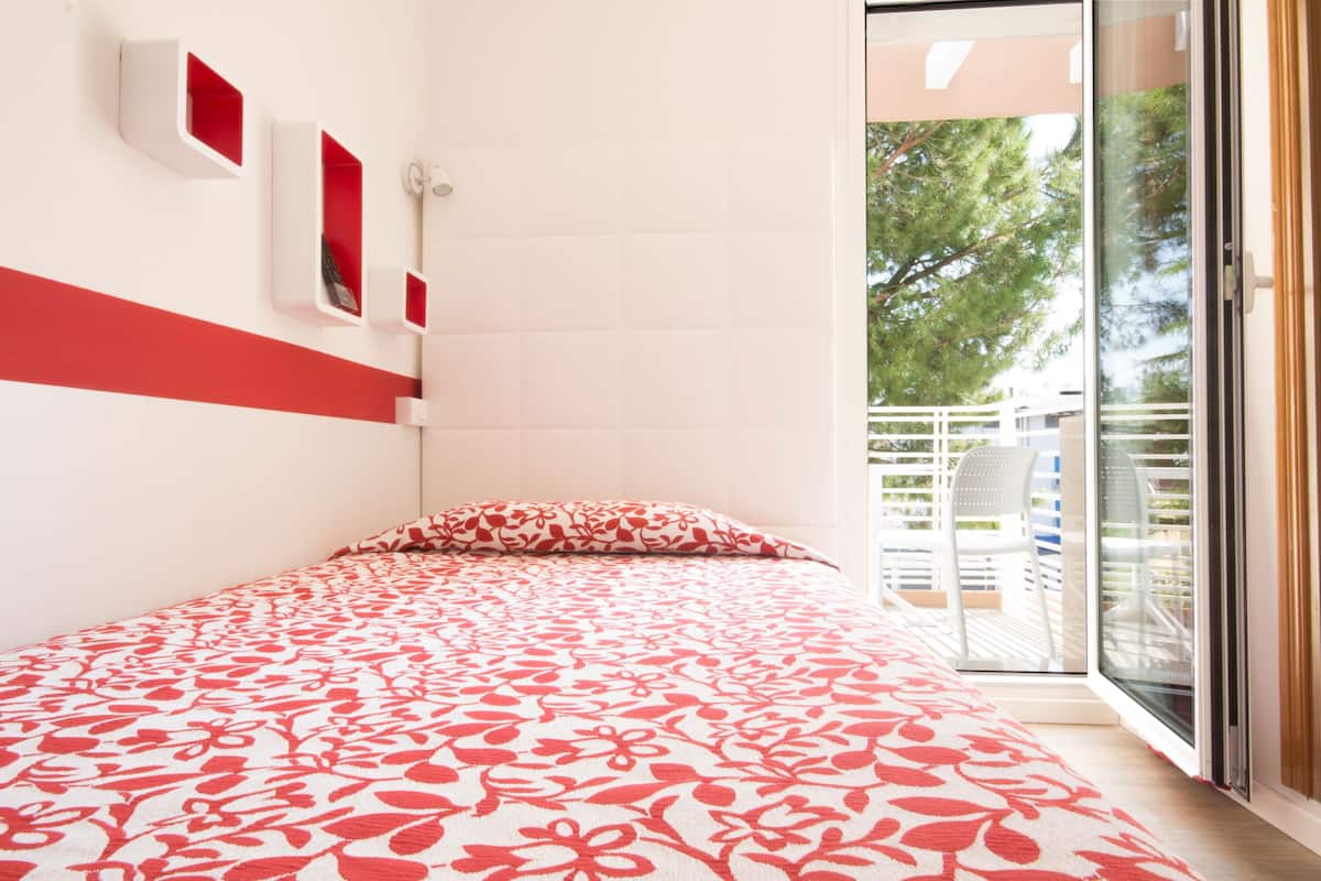 Un dormitorio con cama, colcha roja y blanca estampada, cabecera blanca y puerta corrediza de vidrio que da a un área exterior con una silla y una mesa blancas.