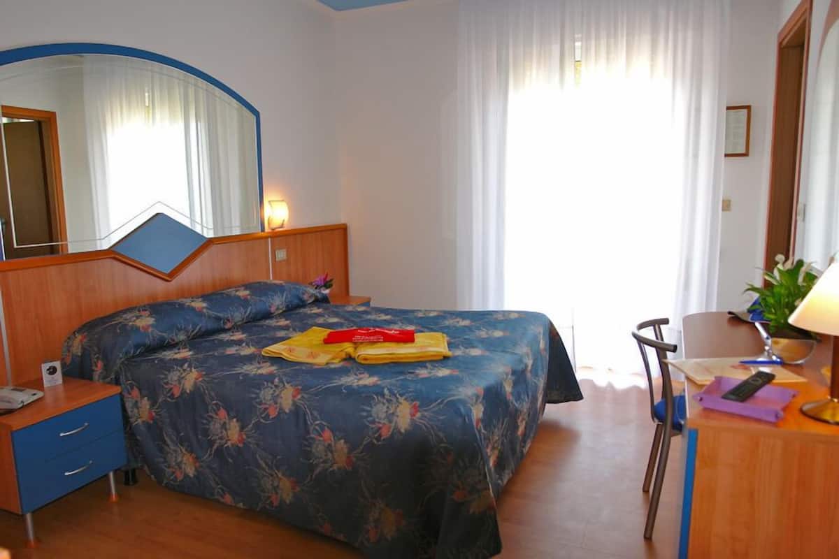 Habitación de hotel con cama, escritorio, silla, espejo y ventana con cortinas.