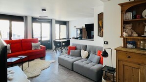 Ein modernes Wohnzimmer mit einem roten Sofa, einem grauen Sofa und einem Esstisch. Im Hintergrund ist eine Küche mit einem Fernseher und einem Bücherregal mit Stofftieren zu sehen.