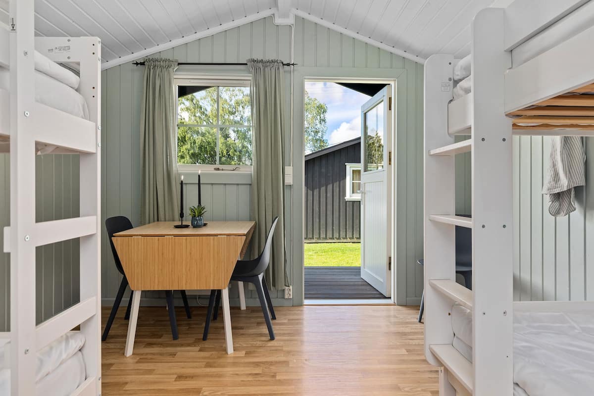 A room with a wooden table and chairs, a bunk bed, and a window with curtains.