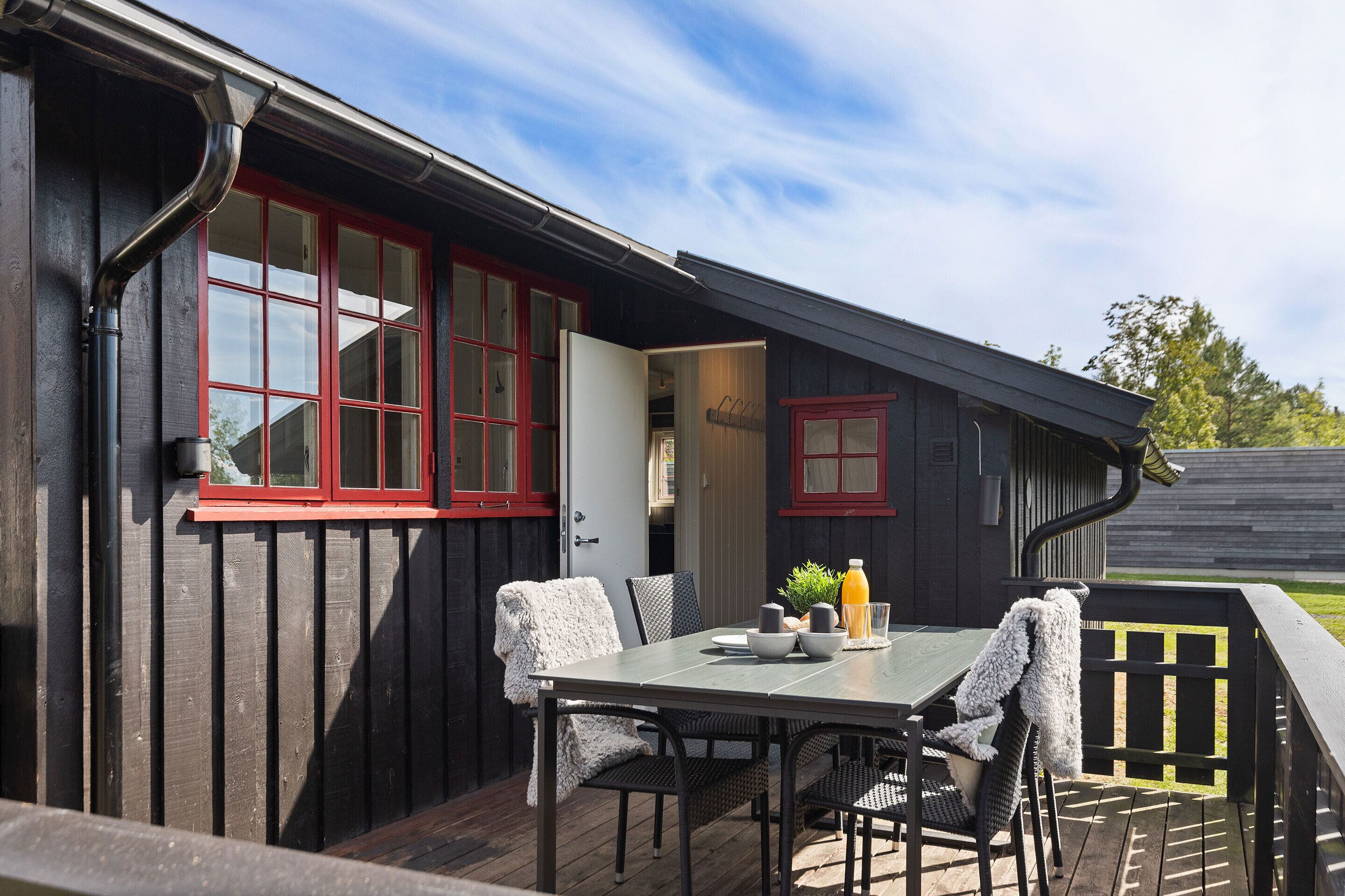 A wooden deck with a table and chairs, a red-framed window, and a door leading to an interior space.