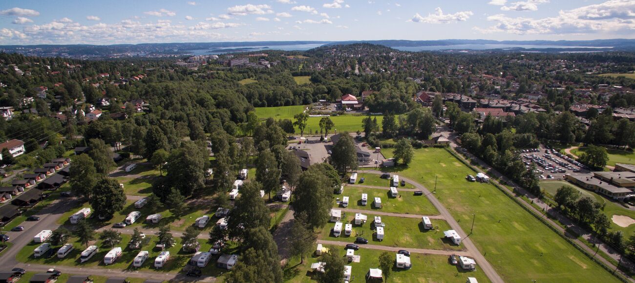 Vista aérea de un camping con vehículos recreativos y tiendas de campaña, rodeado de árboles y zonas residenciales.