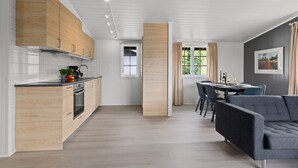 Een moderne keuken met houten kastjes, een zithoek met een grijze bank, en een groot raam met gordijnen.