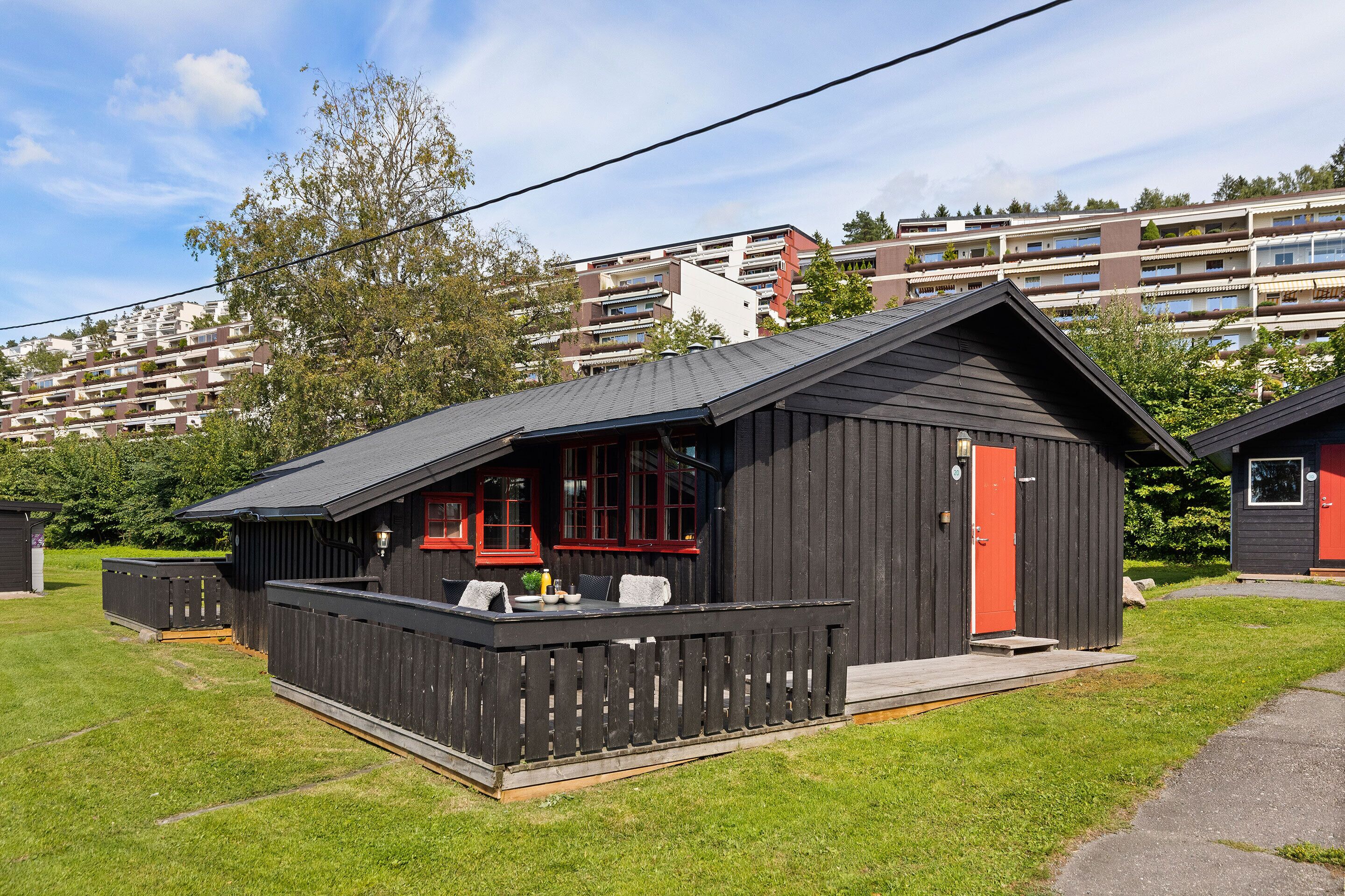 Une petite cabane en bois sombre, avec une porte rouge et un balcon, entourée d’une zone herbeuse et d’un immeuble à plusieurs étages en arrière-plan.