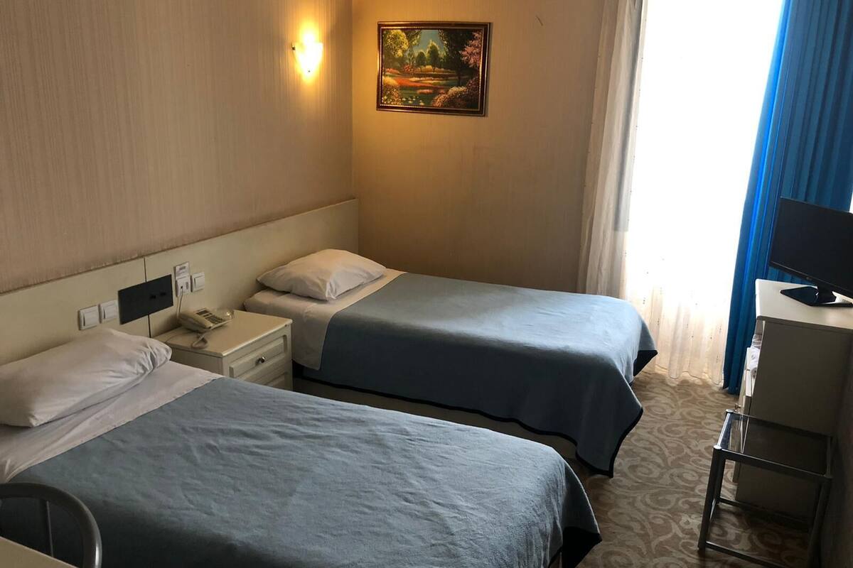 Habitación de hotel con dos camas, un escritorio con computadora, un cuadro en la pared y una ventana con cortinas.