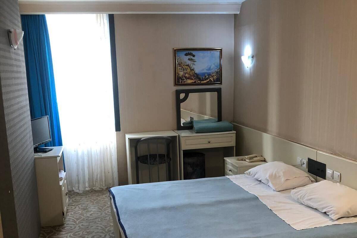 Habitación de hotel con cama, escritorio, silla, pequeño armario, un cuadro en la pared y una ventana con cortinas.