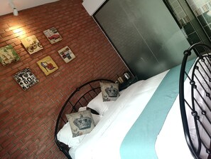 Ein Bett mit Metallgestell, weißer Bettwäsche und Zierkissen. Dahinter befindet sich eine Ziegelwand mit gerahmten Bildern.