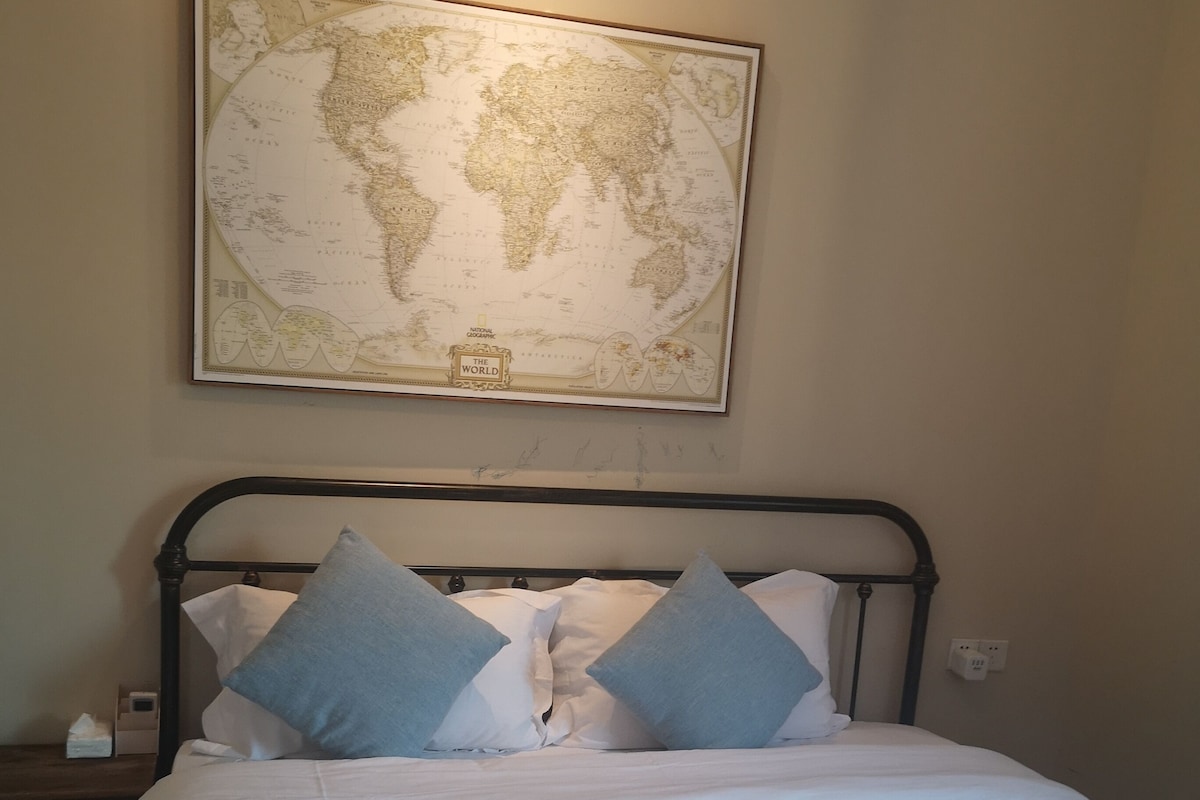 Ein ordentlich bezogenes Bett mit einem gemusterten Überwurf, eine Weltkarte an der Wand darüber und ein Nachttisch mit einer Lampe.