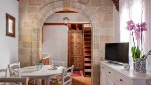 A dining area with a white table and chairs, a TV on a white cabinet, and a stone archway leading to a wooden door. - Acropolis Lindos Home (Rhodes)