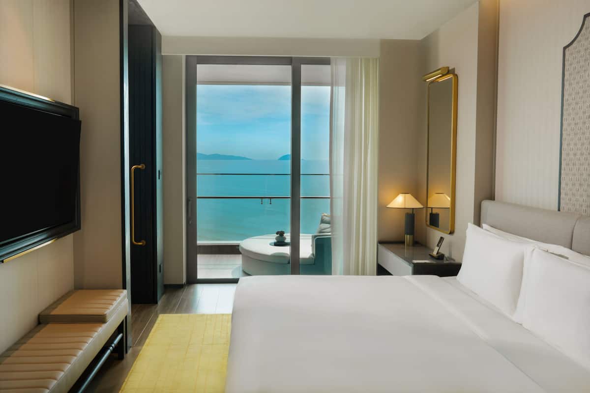 Une chambre d’hôtel comprenant un lit, une télévision, une table de chevet avec une lampe, une chaise et une vue sur la mer, accessible par une porte coulissante.