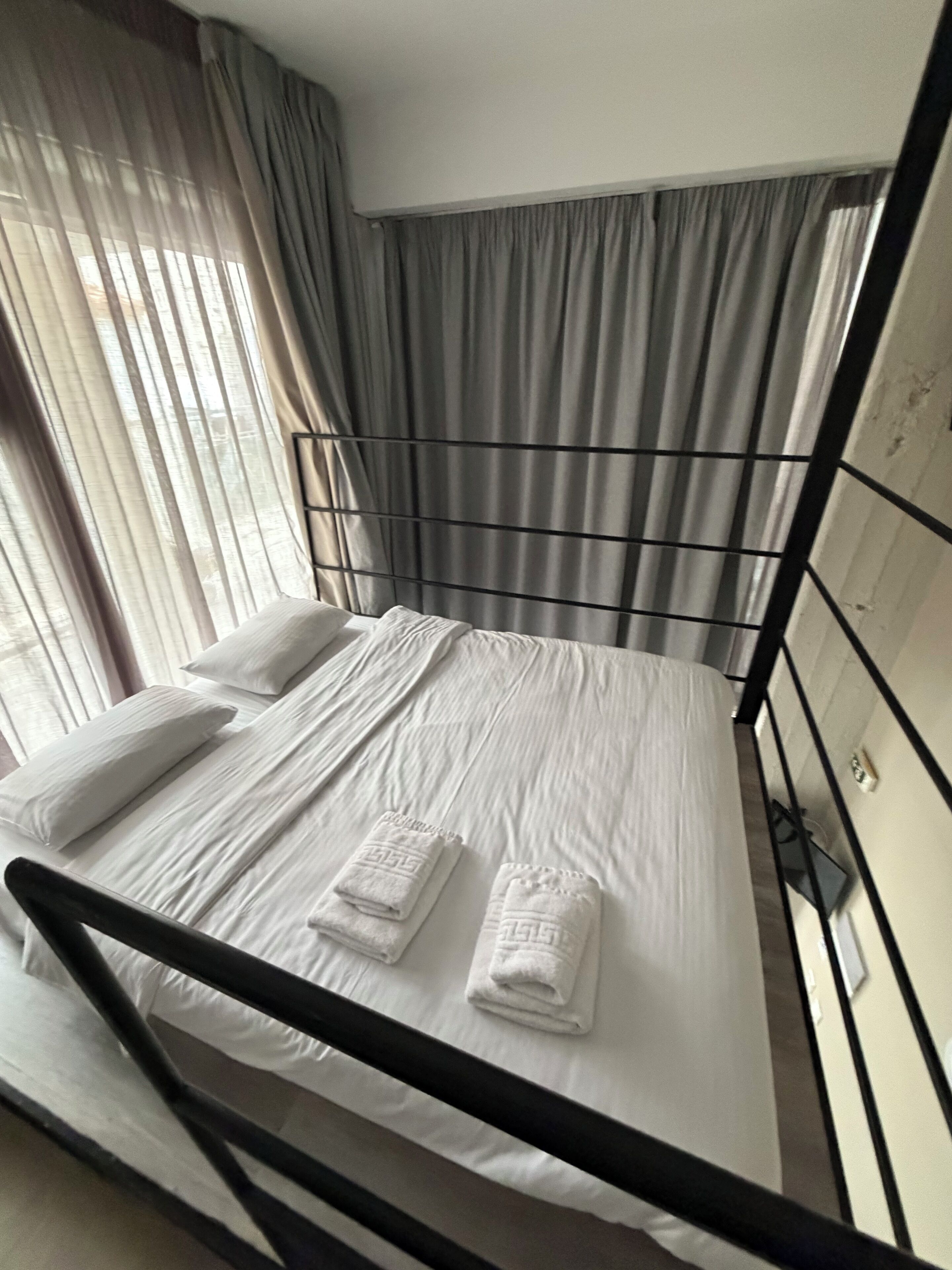 Un lit simple avec du linge de lit blanc et deux serviettes, une fenêtre avec des rideaux légers et une tête de lit en métal.