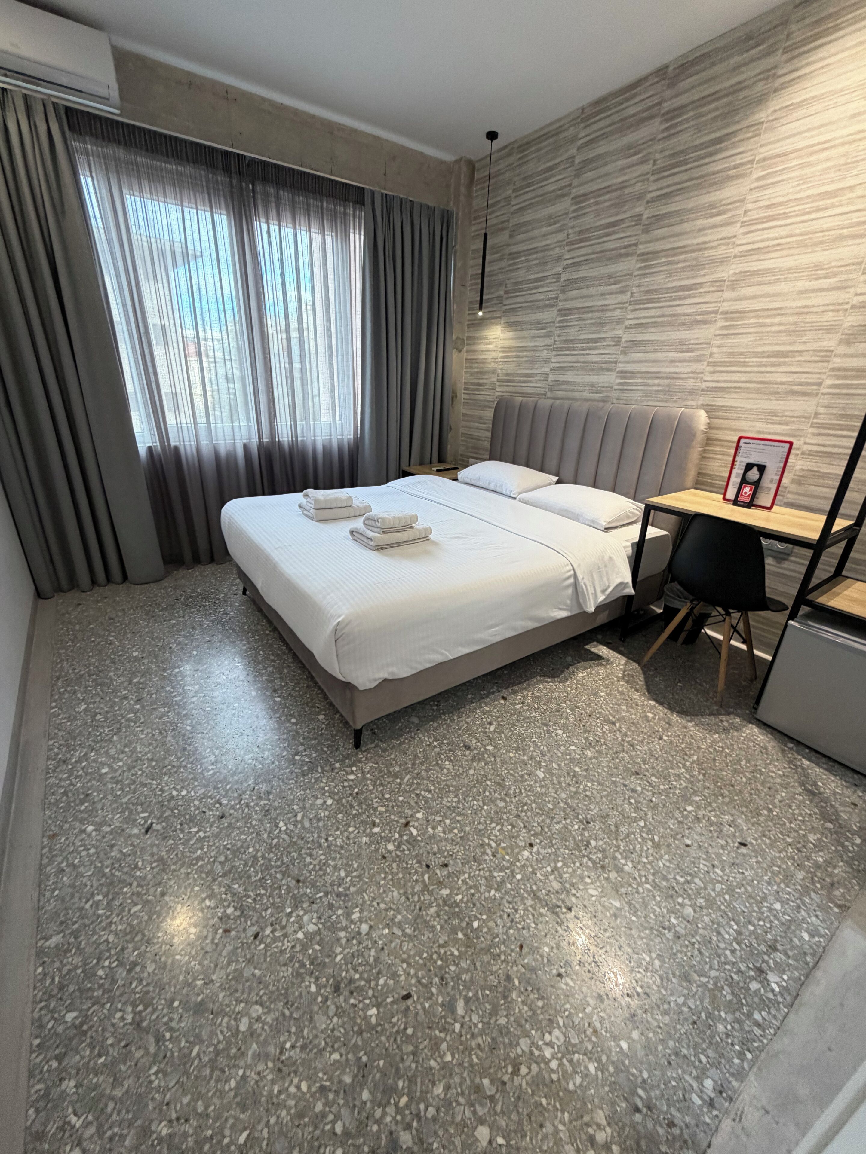 A hotel room with a bed, a small table, a chair, and a wall-mounted air conditioner.