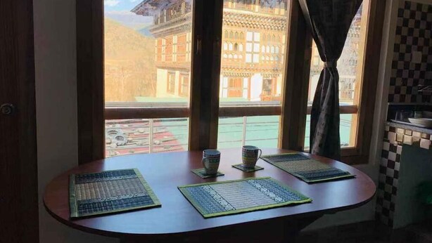 Ein runder Tisch mit zwei Platzsets und zwei Tassen, ein Fenster mit Vorhängen und Blick auf ein Gebäude mit Schachbrettboden.