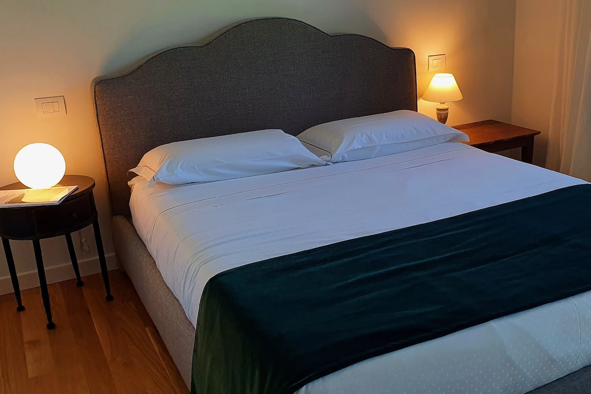 Een net opgemaakt bed met witte lakens en een donkergroene plaid. Twee nachtkastjes, elk met een lamp en een boek. De kamer heeft een eenvoudig, strak ontwerp met een houten vloer.
