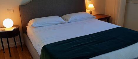 A neatly made bed with white linens and a dark green throw blanket. There are two bedside tables, each with a lamp and a book. The room has a simple, clean design with a wooden floor.