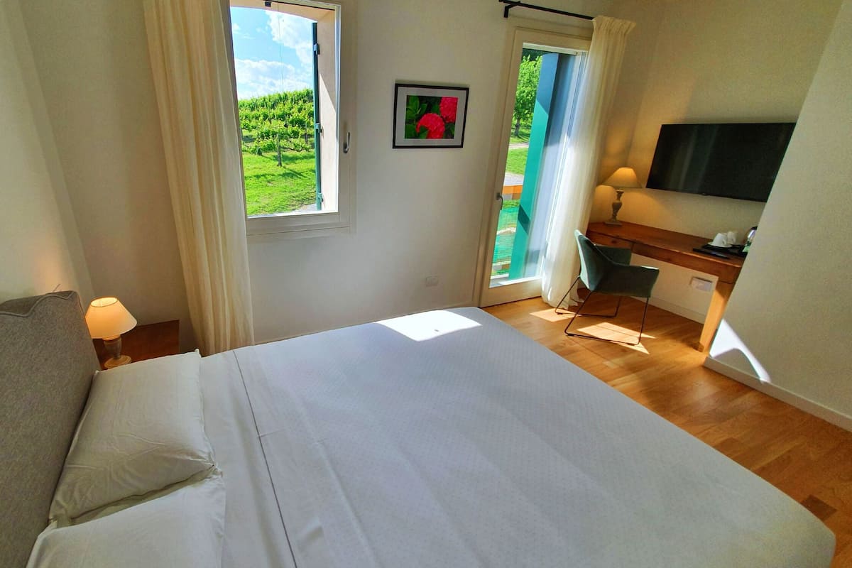 Een slaapkamer met een bed, een bureau, een stoel, een televisie en een raam met uitzicht op groen.