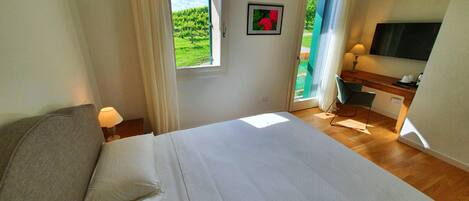 Un dormitorio con cama, escritorio, silla, televisor y ventana con vista a la vegetación.