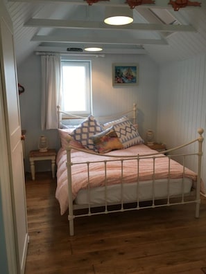 Une chambre avec un lit en métal blanc, une couvre-lit à rayures roses et blanches et des coussins à motifs. Il y a une fenêtre avec des rideaux blancs, une petite table avec une lampe et un tableau au mur.