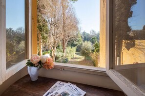 Une fenêtre donnant sur un jardin, un vase rempli de fleurs et un magazine posé sur le rebord de la fenêtre.