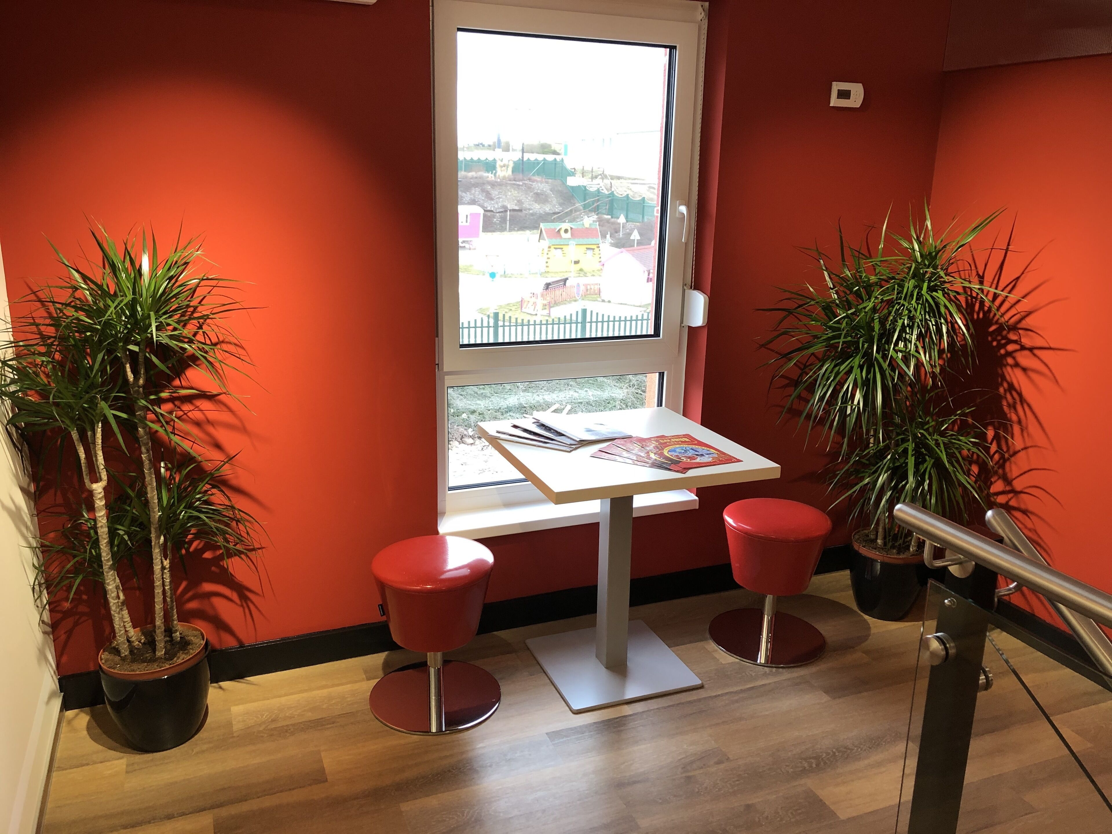 Ein Zimmer mit Fenster, zwei rote Stühle, einem Tisch mit Zeitschriften und zwei Blumentöpfen.