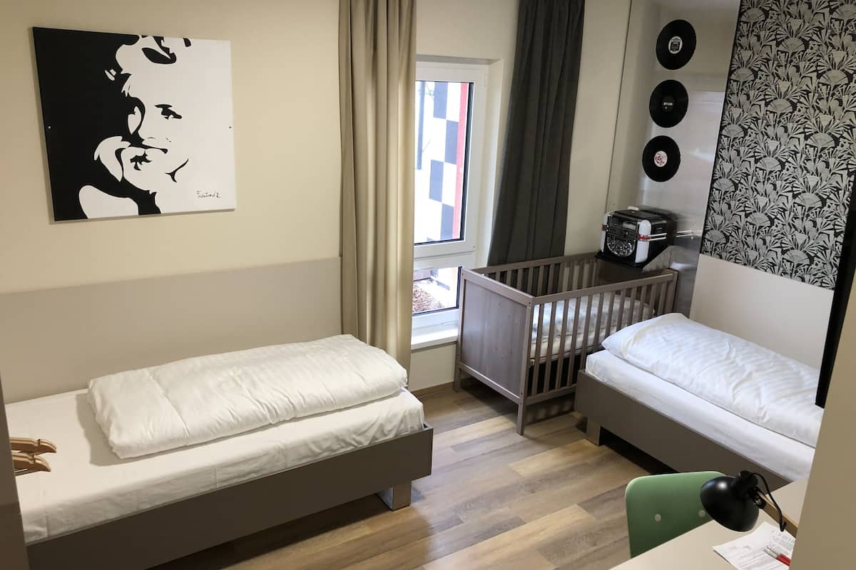 A room with two single beds, a crib, a desk with a chair, a radiator, a window with curtains, and a wall with a black and white artwork.