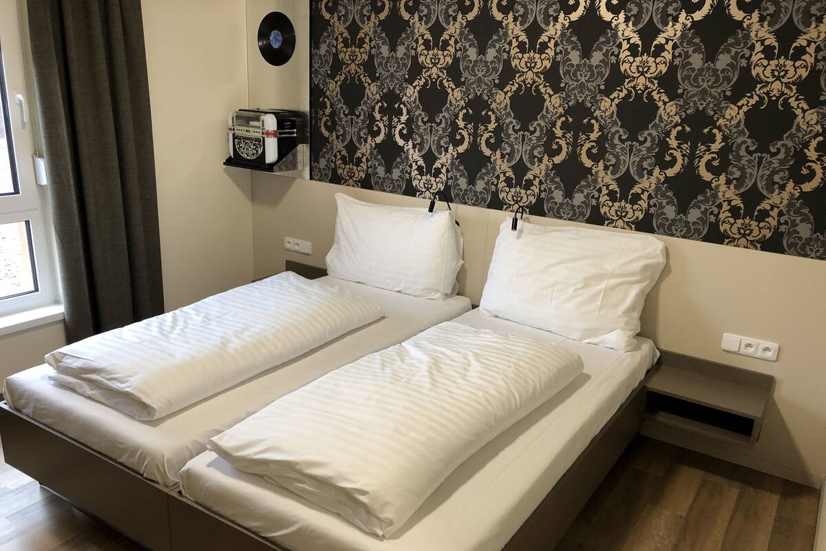 A double bed with white linens, a wall-mounted record player, and a patterned wallpaper.