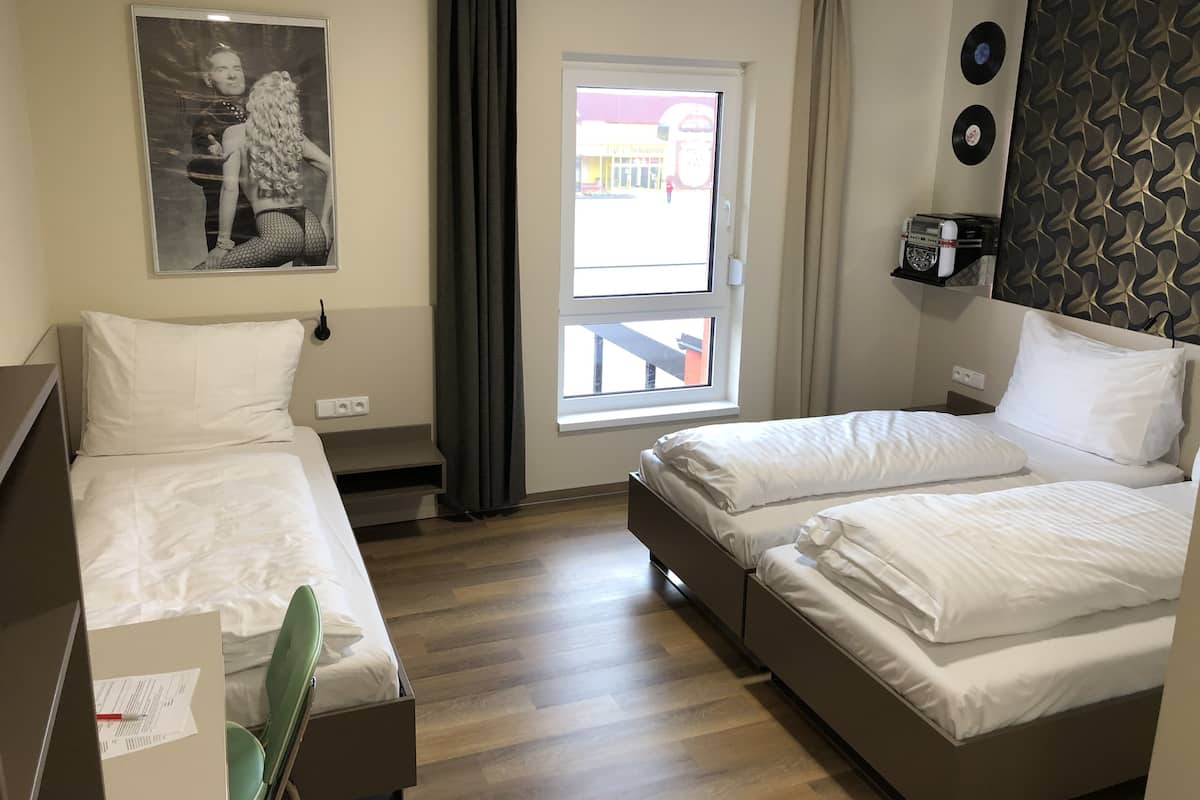 A hotel room with two beds, a window with curtains, a wall-mounted shelf with a radio, and a framed picture on the wall.