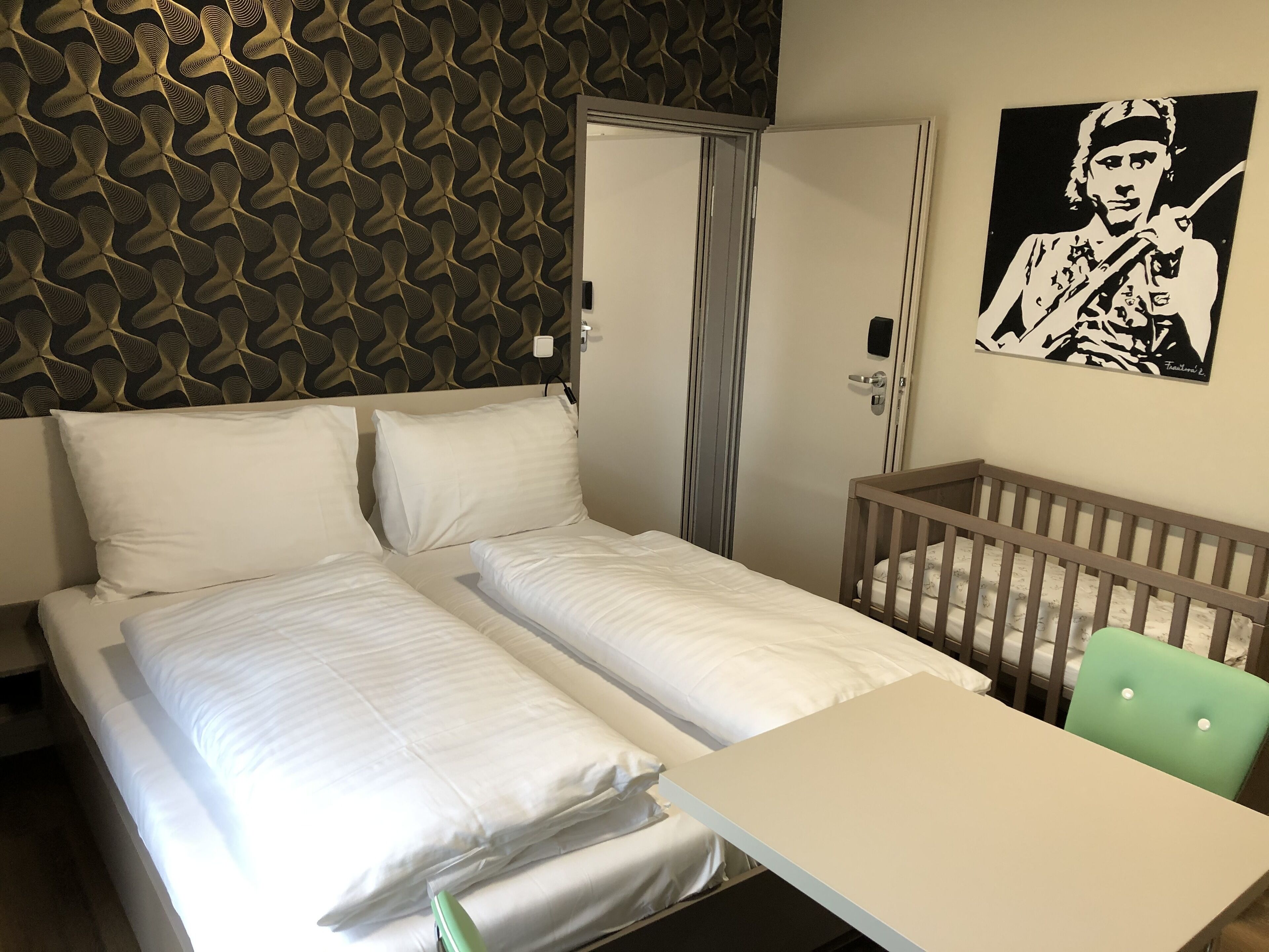 Une chambre d’hôtel avec un lit, un berceau, une table et un mur recouvert d’un papier peint à motifs et orné d’un tableau encadré.
