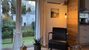 A modern living room with a large window, a black leather chair, a round table with candles and plants, and a kitchen area with wooden cabinets.