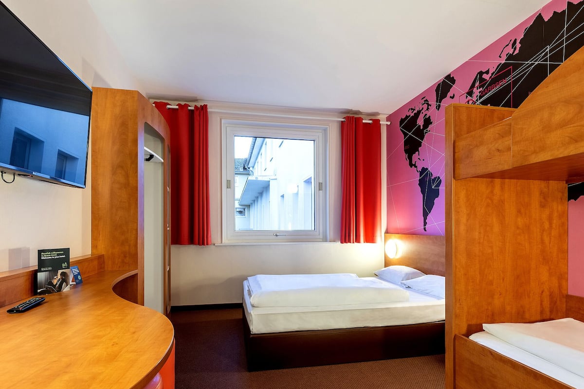 Una habitación de hotel con cama, escritorio, televisión y ventana con cortinas.