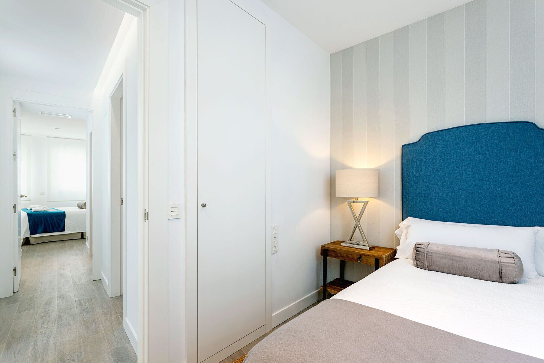 A hotel room with a bed, a nightstand, a lamp, and a door leading to another room.