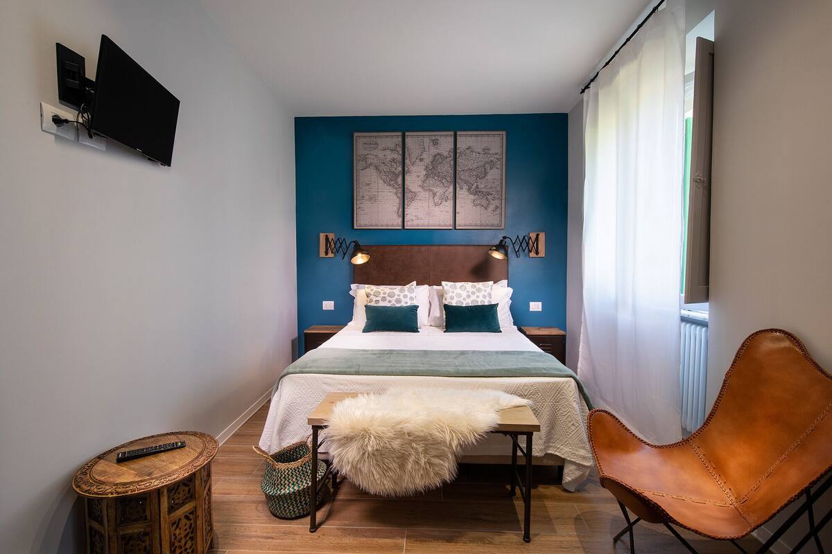 A bedroom with a bed, a chair, a small table, a lamp, a map on the wall, and a window with curtains.