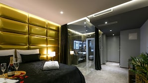 Habitación de hotel con una cama grande, una mesita de noche con una botella y vasos, y un baño visible a través de una puerta de cristal.