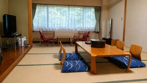 Une pièce de style japonais traditionnel, avec un sol recouvert de tatamis, une table à manger en bois, des chaises rouges, un téléviseur et une fenêtre ornée de rideaux légers.