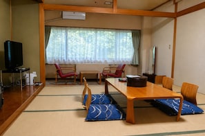Ein traditionell gestalteter japanischer Raum mit Tatami-Bodenbelag, einem Holztisch, roten Stühlen, einem Fernseher und einem Fenster mit durchsichtigen Vorhängen.