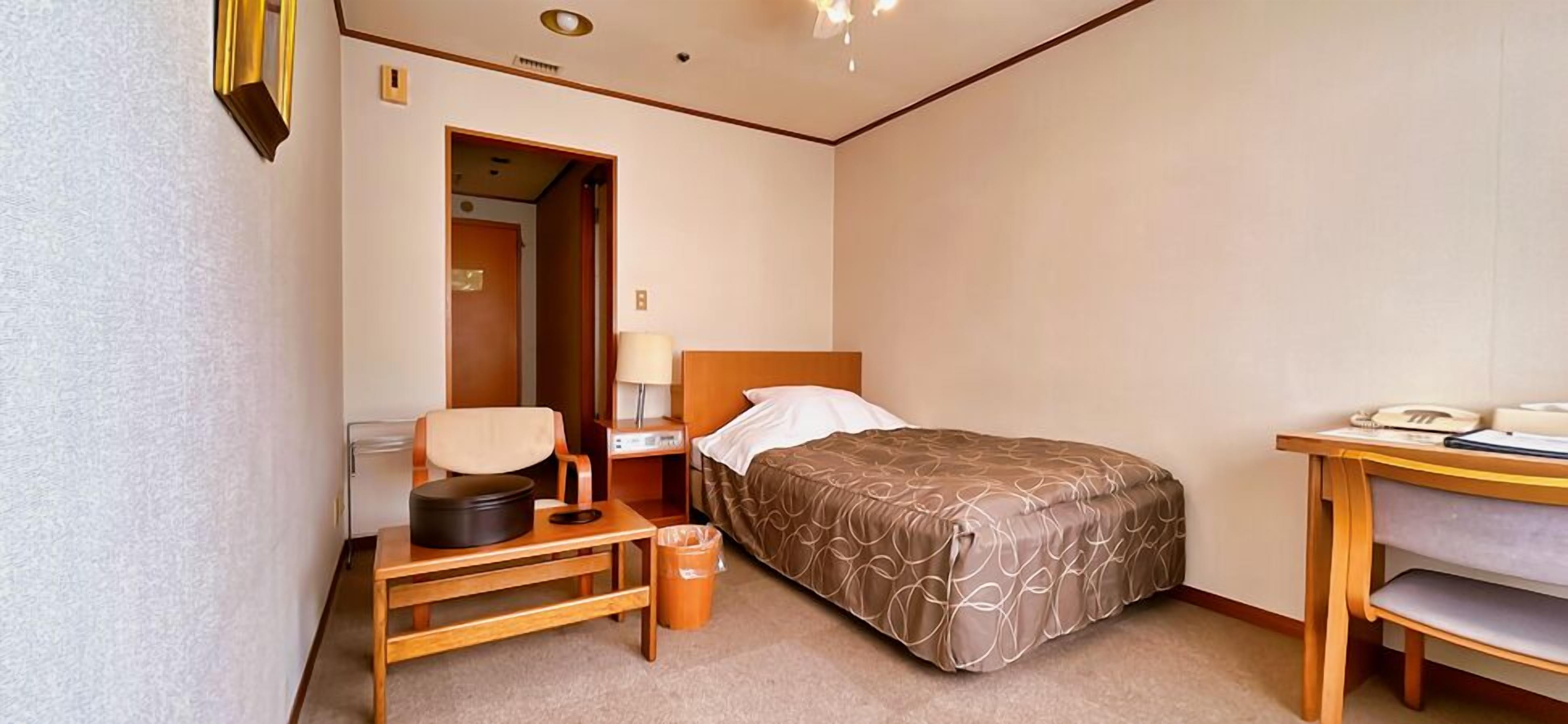 Habitación de hotel con cama, mesita de noche, silla, lámpara y escritorio con teléfono.