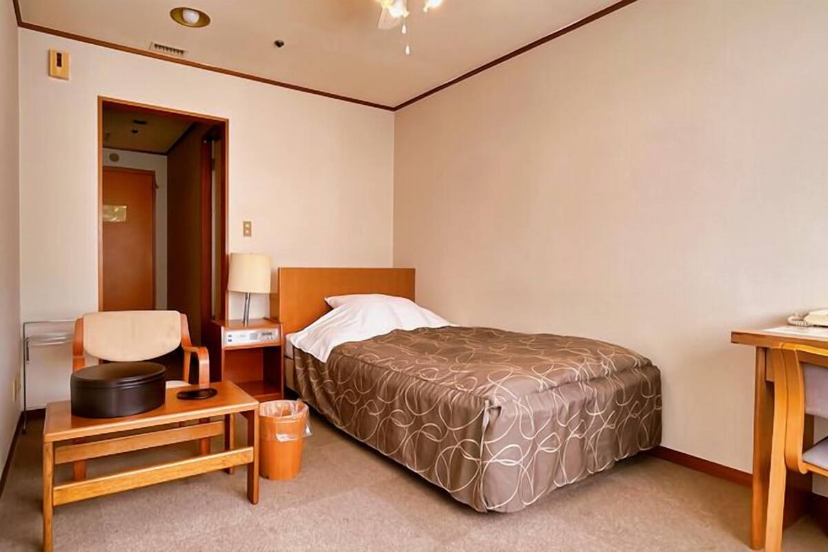 Habitación de hotel con cama, mesita de noche, silla, lámpara y escritorio con teléfono.