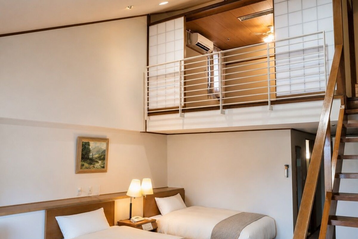 A hotel room with two beds, a small desk with a lamp, a painting on the wall, and a staircase leading to an upper level.