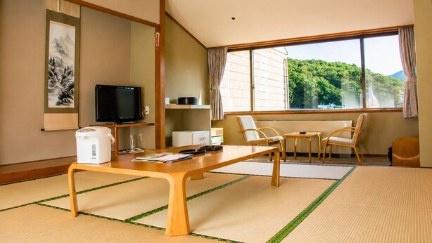 Une pièce de style japonais traditionnel, avec un sol recouvert de tatamis, une table basse, un téléviseur et une vue sur la verdure environnante.