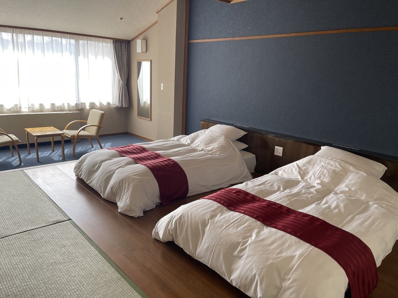 Habitación de hotel con dos camas, zona de estar con sillas, un ventanal grande con cortinas y suelo de madera.